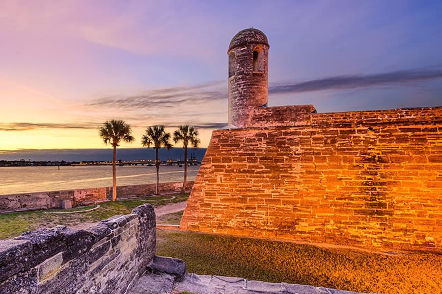 A stone fort tower stands beside a calm waterway at sunset, framed by palm trees.