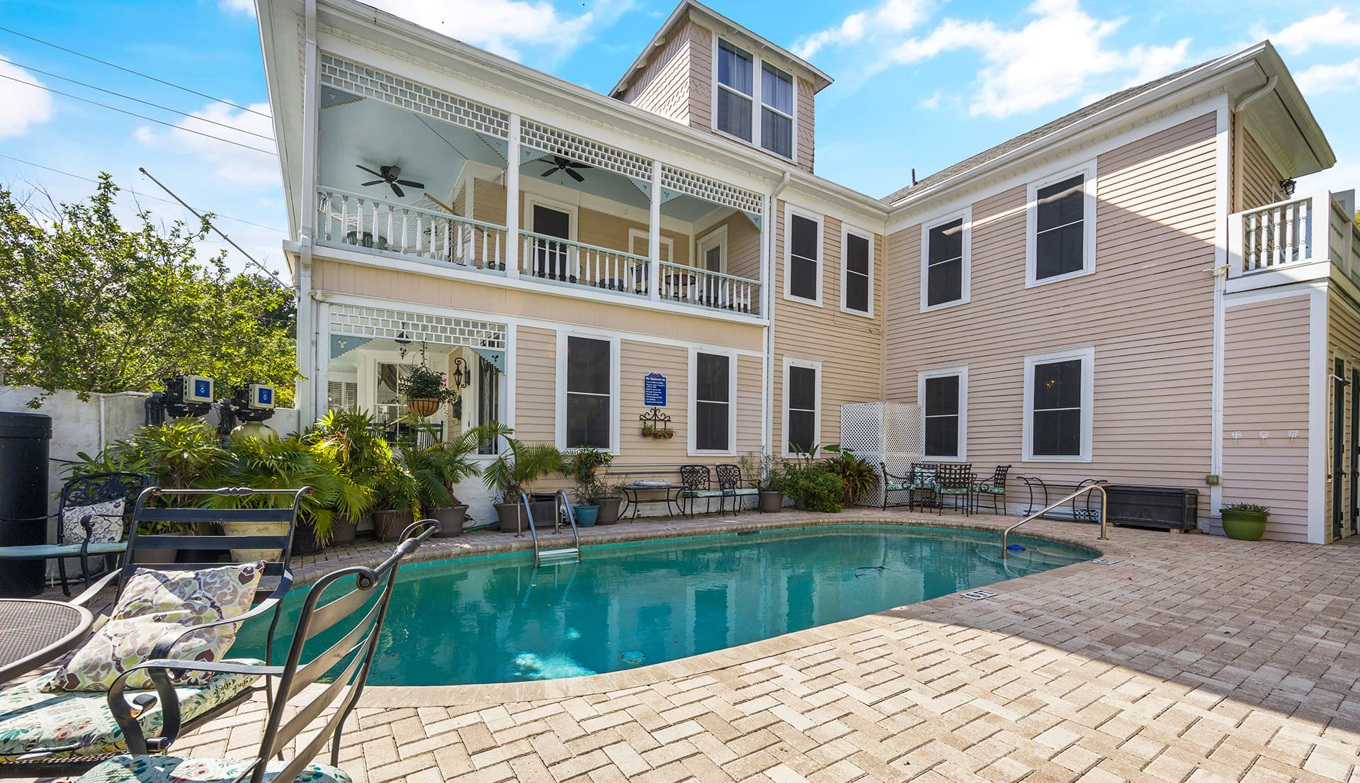 A sunny pool area surrounded by a charming two-story house and lush greenery.