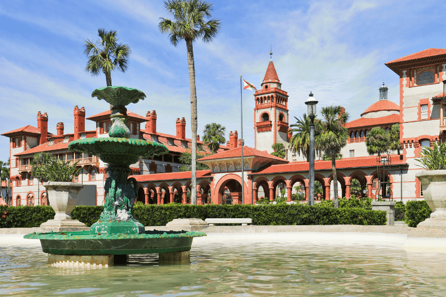 A historic building with red roofs and a clock tower beside a fountain and palm trees.