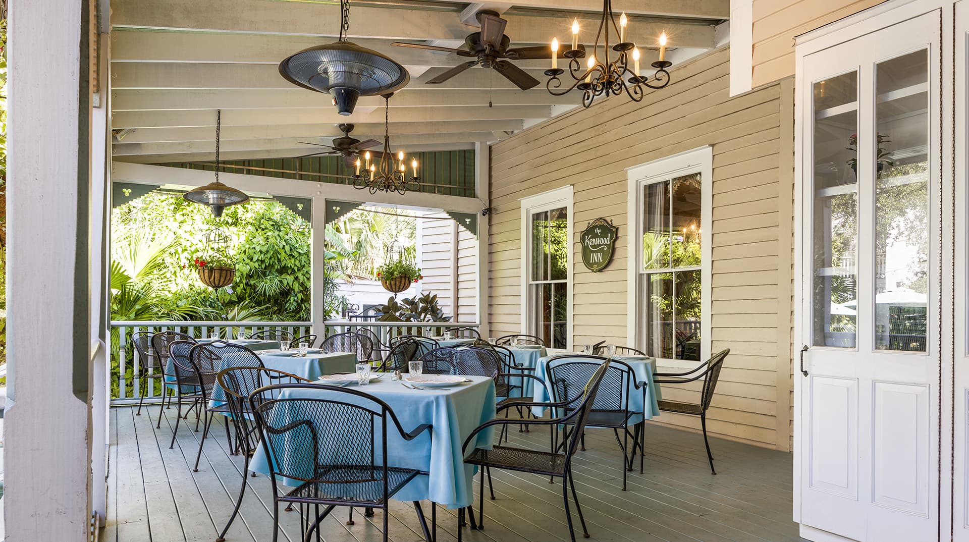 A porch dining area with tables set in light blue cloth and black chairs, surrounded by greenery.