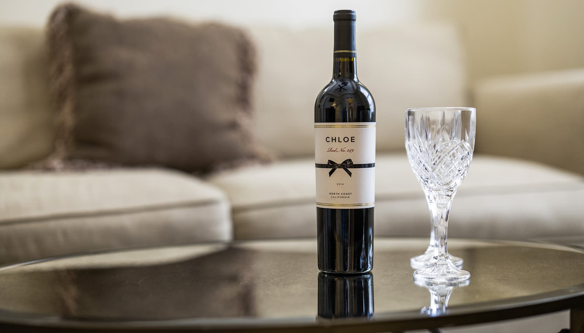 A bottle of Chloe red wine stands next to a crystal glass on a dark coffee table, with a soft sofa in the background.