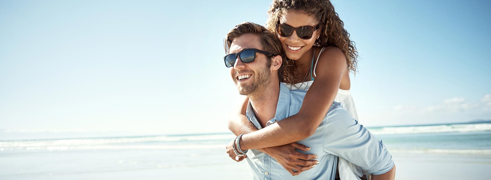 A man gives a woman a piggyback ride on the beach, both smiling and wearing sunglasses.