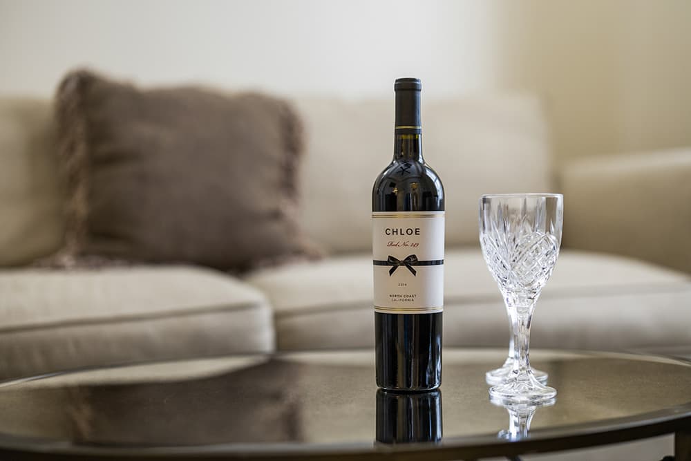 A bottle of Chloe wine stands next to a crystal wine glass on a glass table in a cozy living room setting.