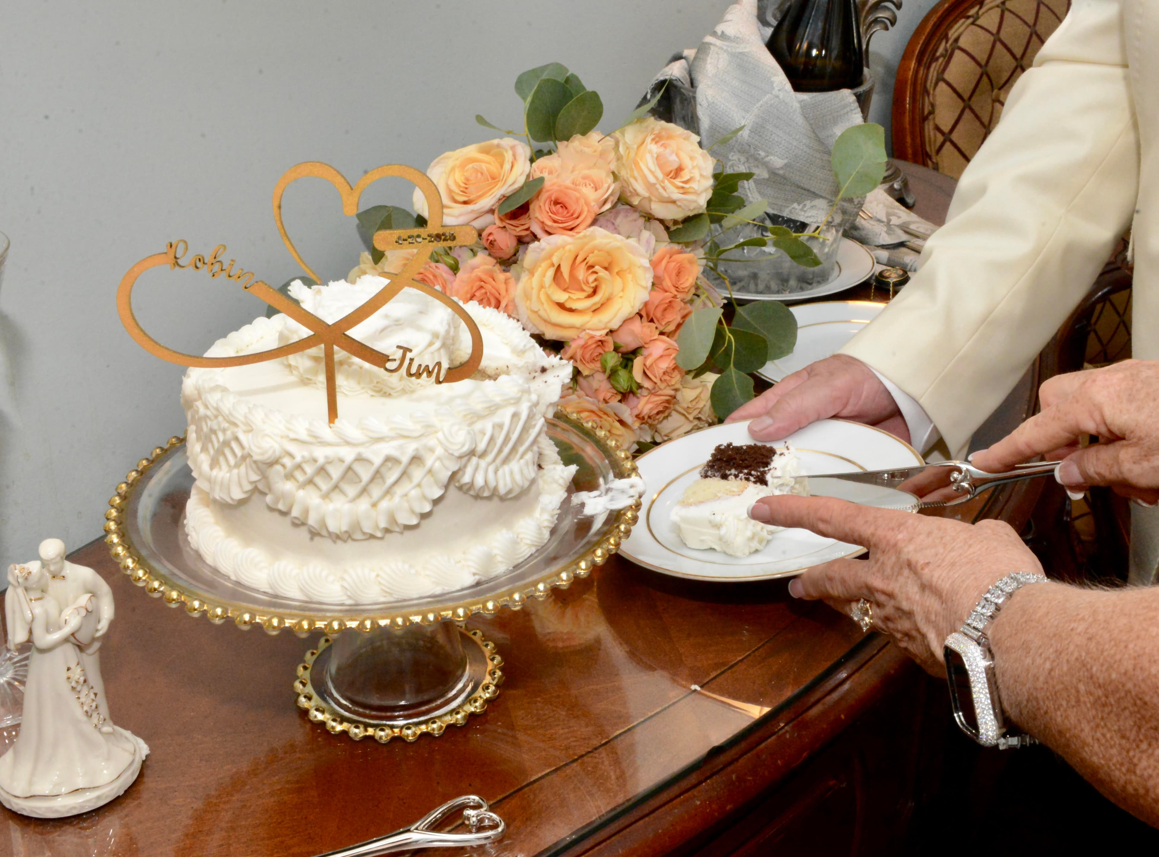 A hand serves a slice of decorated wedding cake beside a bouquet of roses and a couple figurine.