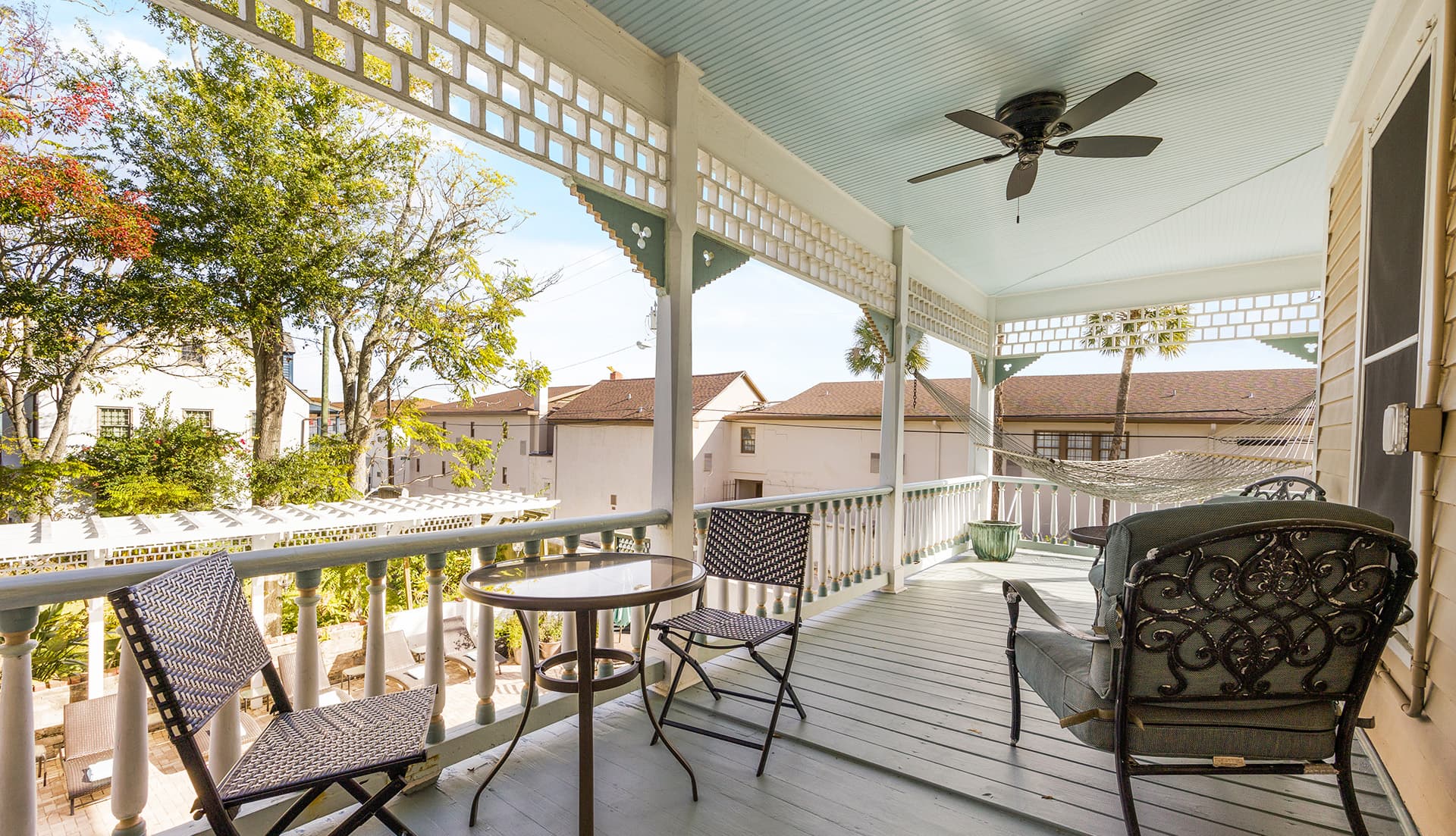 A cozy porch with seating and a ceiling fan, surrounded by greenery and buildings.